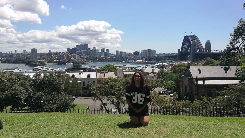 Vivir en Australia chica morena de rodillas en el césped y de fondo la ciudad de Sydney con el cielo despejado