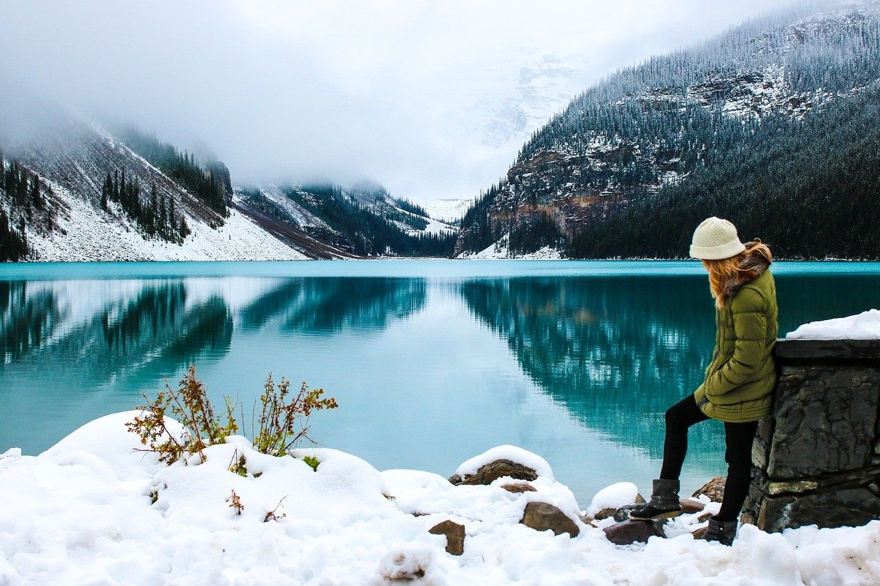 Vivir en Canadá chica abrigada encima de la nieva al lado de un lago rodeado de montañas