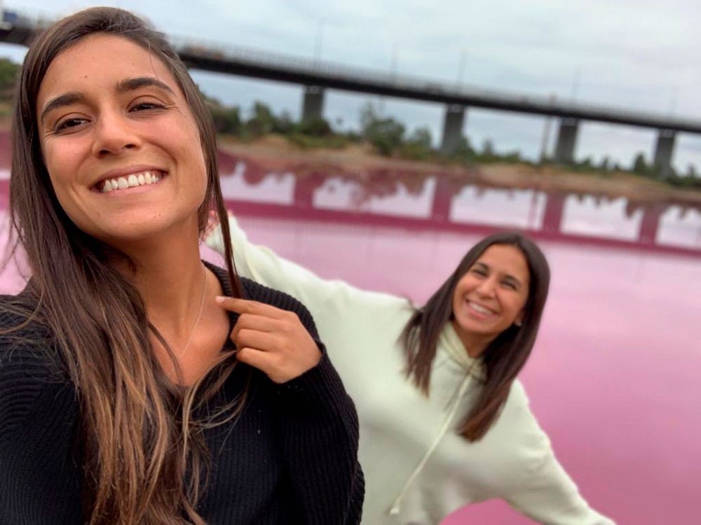 dos chicas morenas sonriendo a cámara y detrás un lago rosa