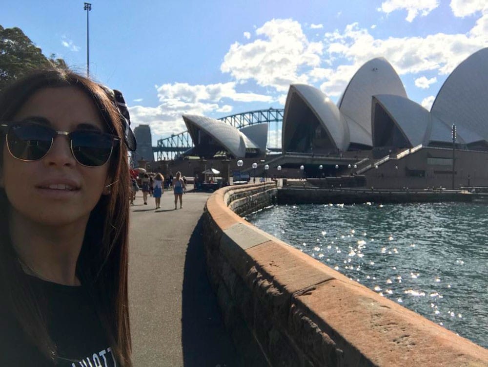 chica morena con gafas sonriendo a cámara y detrás la Opera House de Sydney