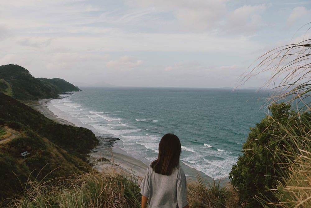 pagina nueva zelanda chica morena de espaldas mirando al mar y rodeada de vegetación