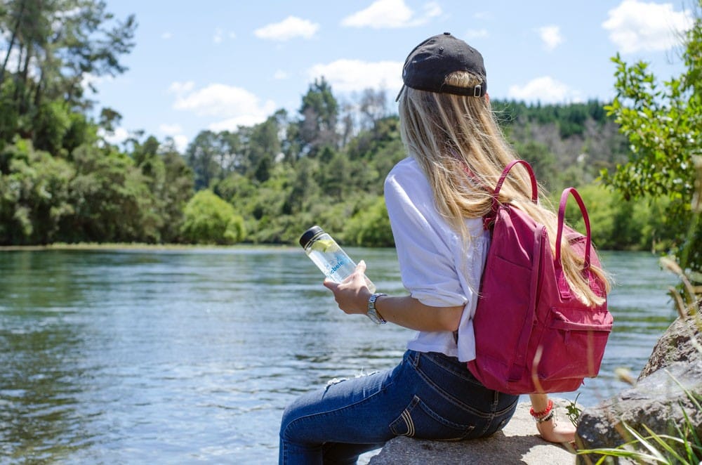 cuánto cuesta Nueva Zelanda chica rubia de espaldas sentada en una roca de un lago