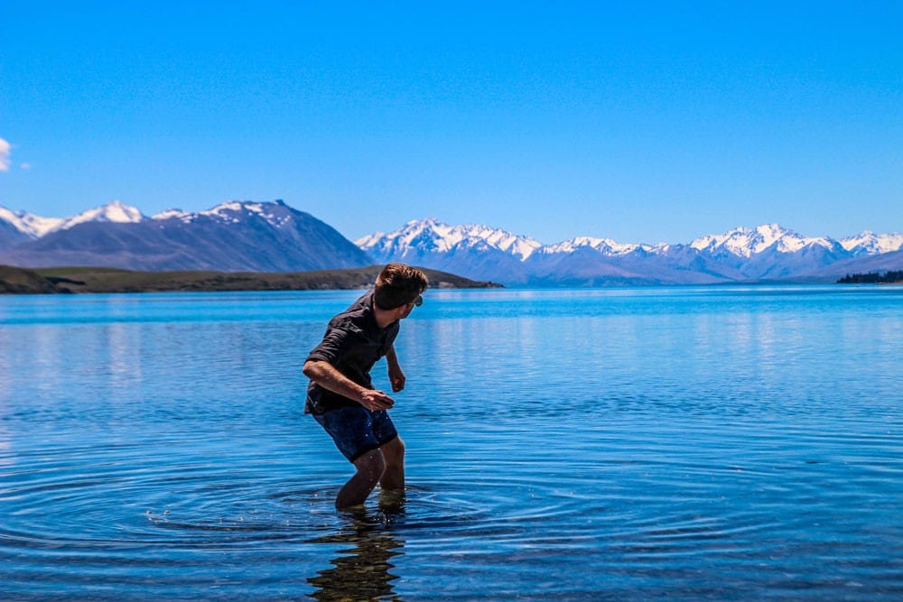 estudiar en nueva zelanda chico de espaldas tirando una piedra en un lago y detrás montañas