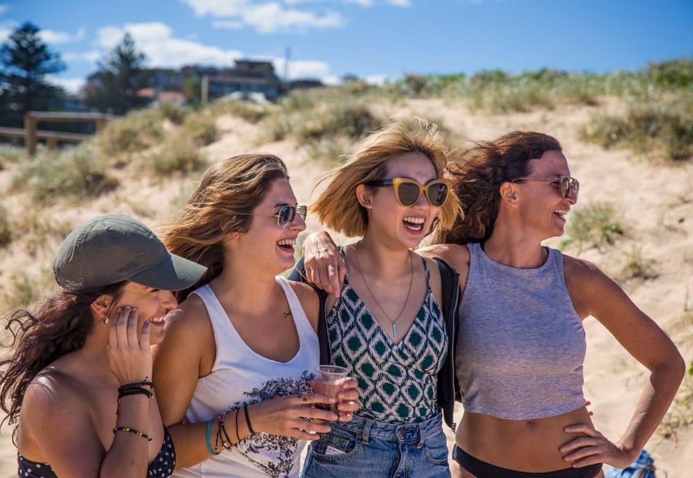 cuatro chicas con gafas de sol sonriendo y posando para la fotografía. Están encima de la arena y detrás el cielo despejado