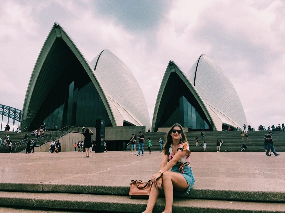 chica morena sentada en unas escaleras y detrás la Ópera de Sydney. Ella lleva gafas de sol y shorts tejanos