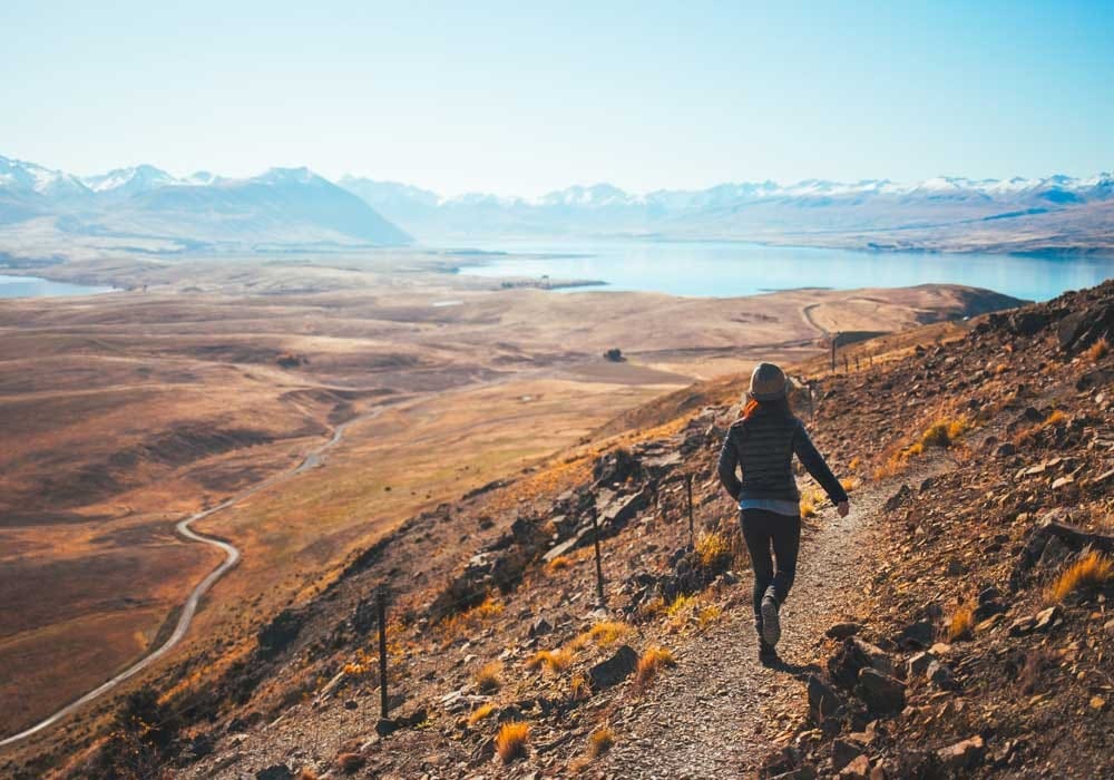 cuánto cuesta vivir en Nueva Zelanda chica de espaldas andando por una montaña y al fondo un lago