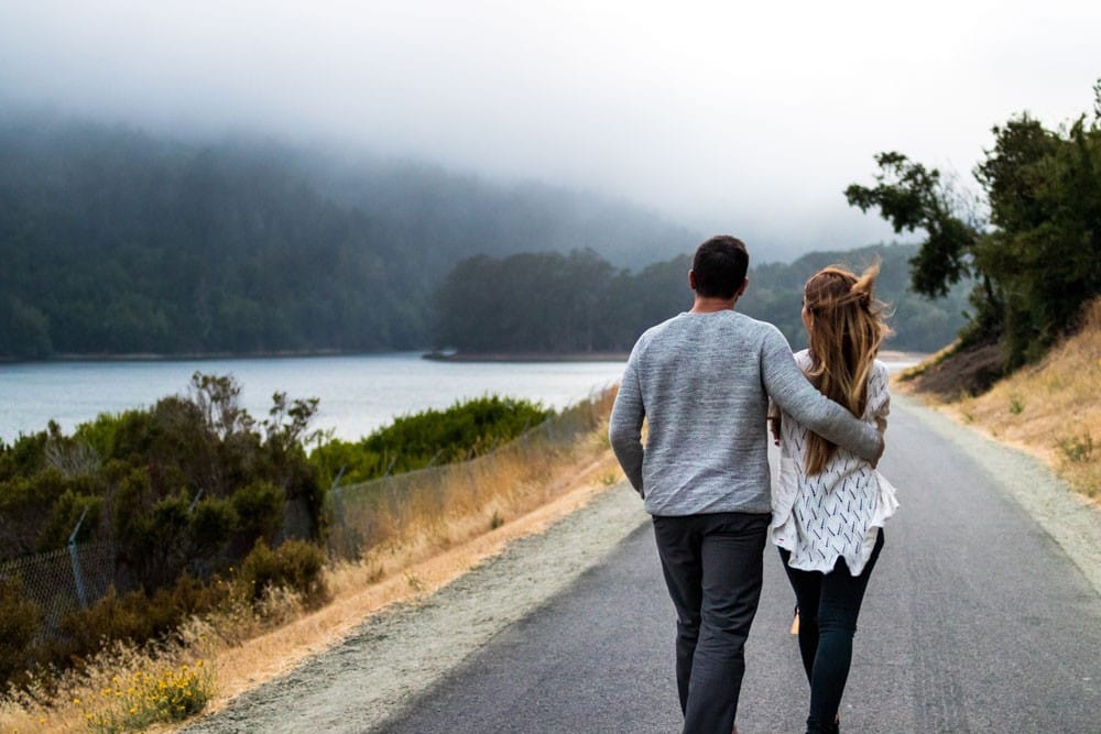 Visado de pareja pareja andando por una carretera y delante el paisaje de una montaña con el lago
