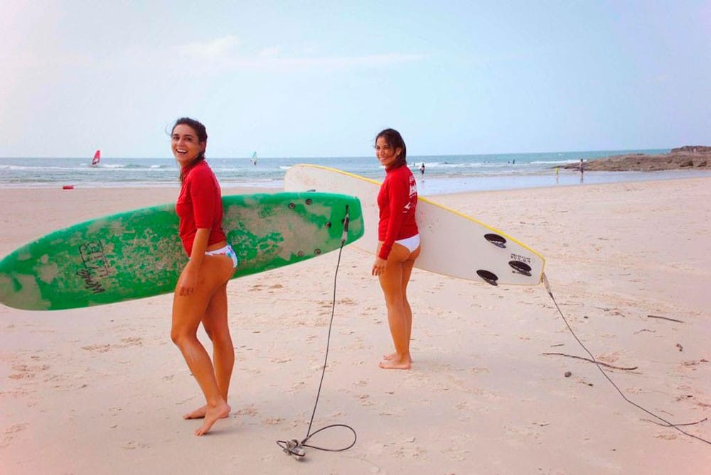 dos chicas con neopreno rojo y una tabla de surf sonriendo a cámara