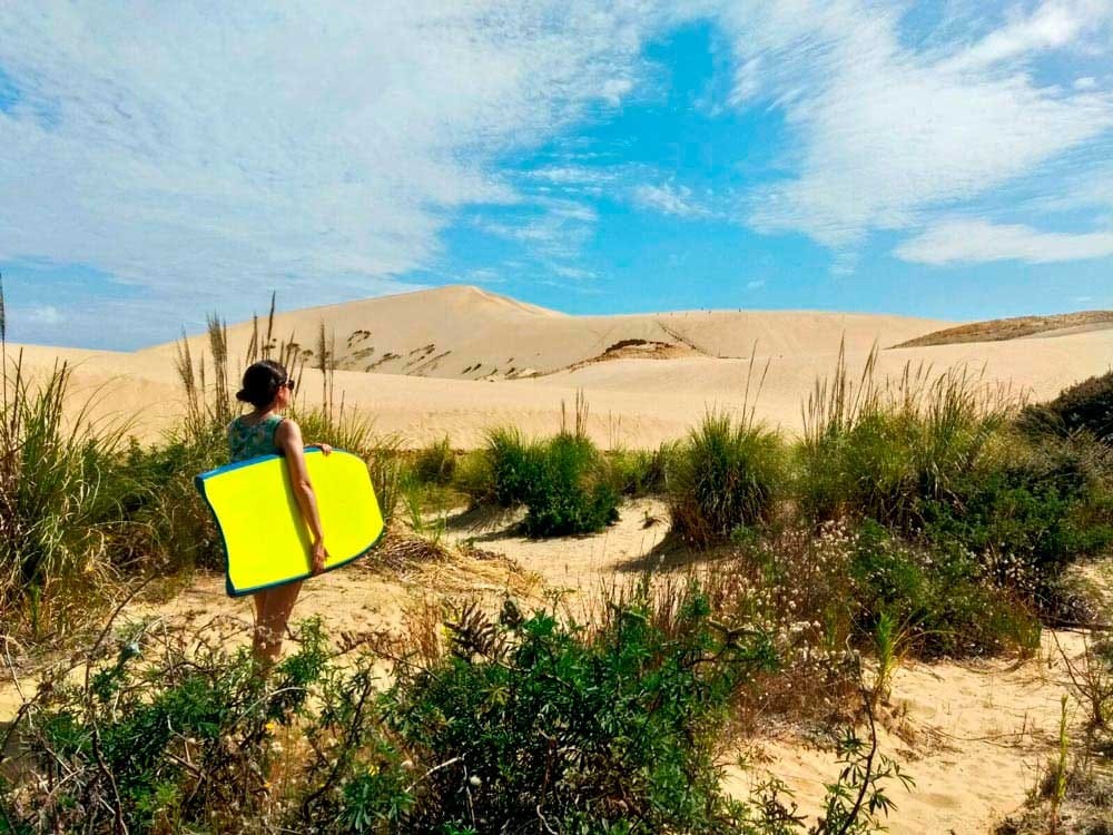 Residencia permanente chica de espaldas con una tabla de surf amarilla andando por una duna
