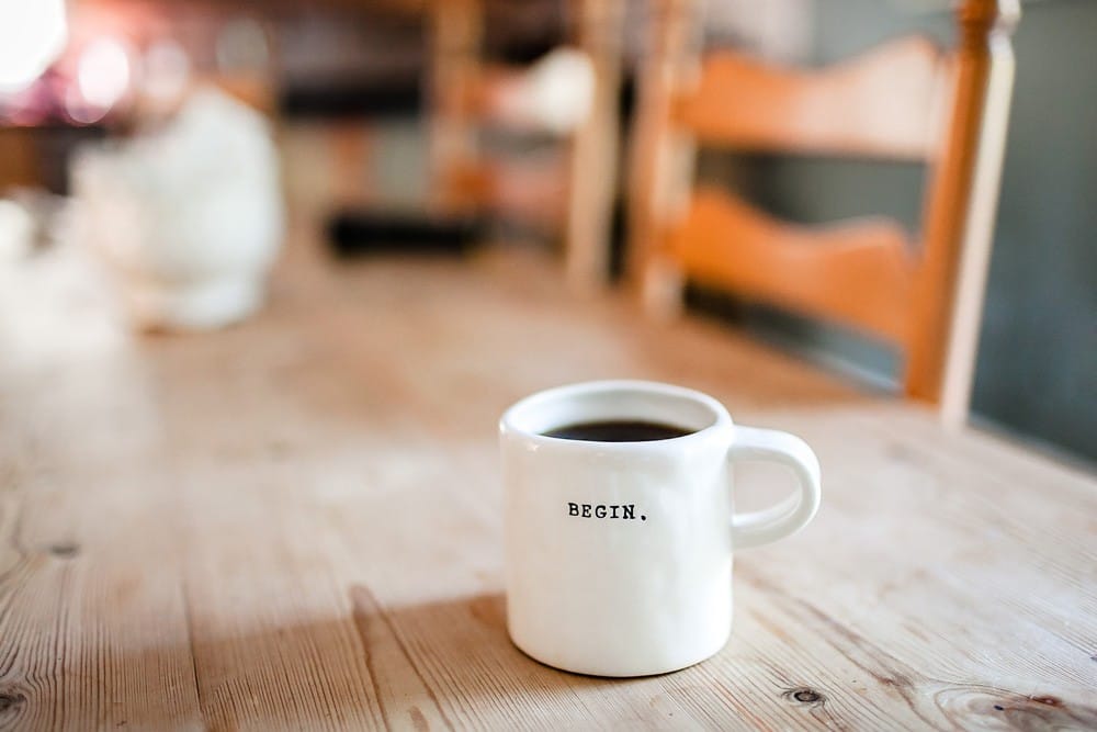 vivir en canadá fotografía de una taza blanca con café encima de una mesa de madera. En la taza pone "begin"