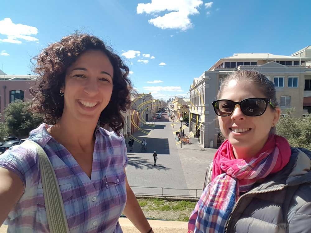 vivir en Australia dos chicas morenas sonrientes mirando a cámara y detrás edificios
